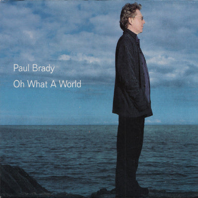Album cover with a person standing by the sea and 'Paul Brady Oh What A World' text.